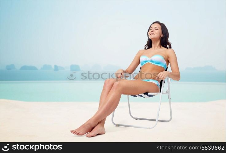 people, tanning, summer and beach concept - happy young woman in bikini swimsuit sunbathing on folding chair over infinity edge pool at hotel resort background