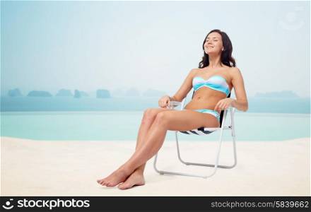 people, tanning, summer and beach concept - happy young woman in bikini swimsuit sunbathing on folding chair over infinity edge pool at hotel resort background