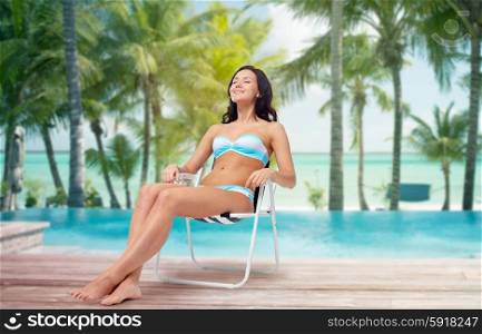 people, tanning, summer and beach concept - happy young woman in bikini swimsuit sunbathing on folding chair over tropical beach with palm trees and pool at hotel resort background