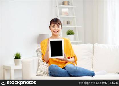 people, technology, advertisement and leisure concept - happy young asian woman sitting on sofa and showing tablet pc computer blank black screen at home