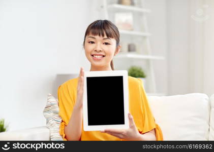 people, technology, advertisement and leisure concept - happy young asian woman sitting on sofa and showing tablet pc computer blank black screen at home