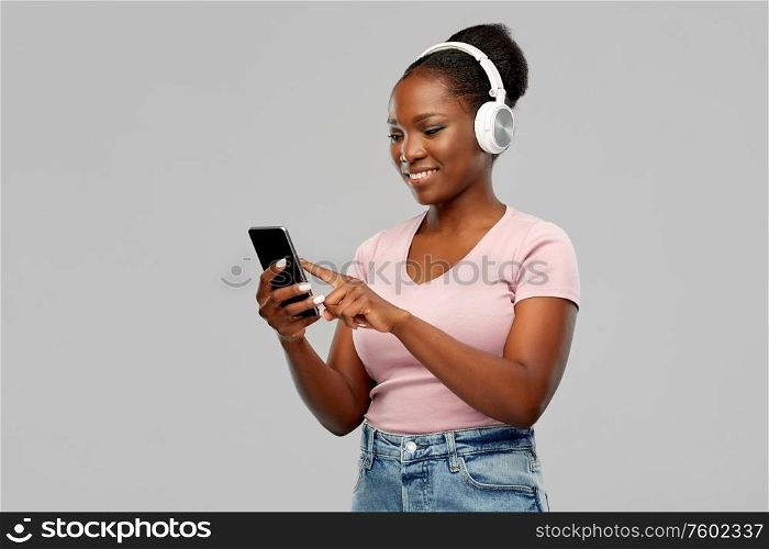 people, technology and audio equipment concept - happy african american young woman in headphones listening to music on smartphone over grey background. african woman in headphones listening to music