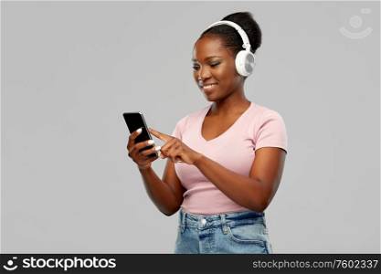 people, technology and audio equipment concept - happy african american young woman in headphones listening to music on smartphone over grey background. african woman in headphones listening to music