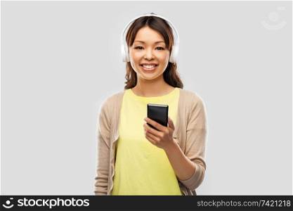 people, technology and audio equipment concept - happy asian young woman in headphones listening to music on smartphone over grey background. asian woman in headphones listening to music