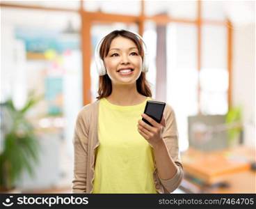 people, technology and audio equipment concept - happy asian young woman in headphones listening to music on smartphone over office background. asian woman in headphones listening to music