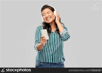 people, technology and audio equipment concept - happy smiling asian young woman in headphones listening to music on smartphone over grey background. happy asian woman in headphones listening to music