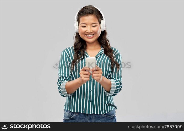 people, technology and audio equipment concept - happy smiling asian young woman in headphones listening to music on smartphone over grey background. happy asian woman in headphones listening to music