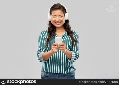 people, technology and audio equipment concept - happy smiling asian young woman in headphones listening to music on smartphone over grey background. happy asian woman in headphones listening to music