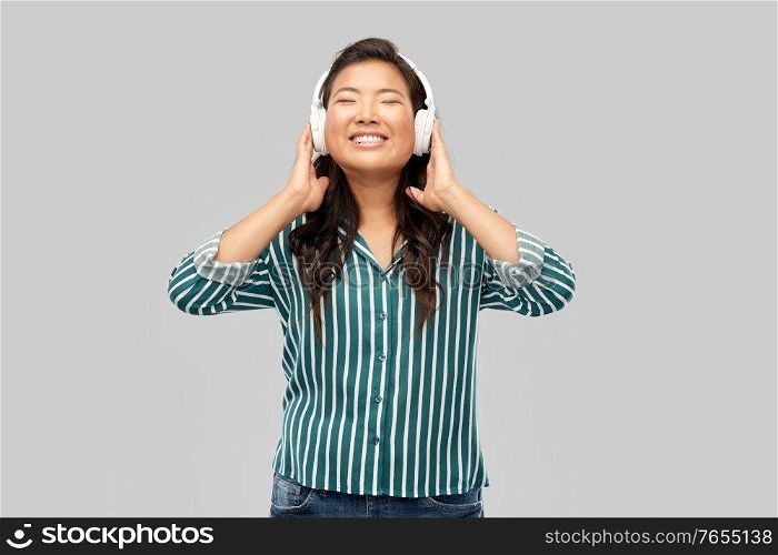 people, technology and audio equipment concept - happy young asian woman in headphones listening to music over grey background. asian woman in headphones listening to music