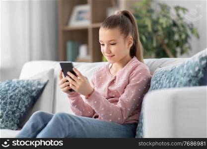 people, technology and childhood concept - happy smiling little girl with smartphone sitting on sofa at home. happy smiling little girl with smartphone at home