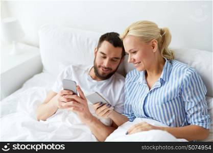 people, technology and communication concept - happy couple with smartphones in bed at home. happy couple with smartphones in bed at home