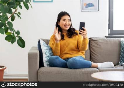 people, technology and communication concept - happy smiling asian young woman in yellow sweater sitting on sofa and having video call on smartphone at home. woman with smartphone having video call at home