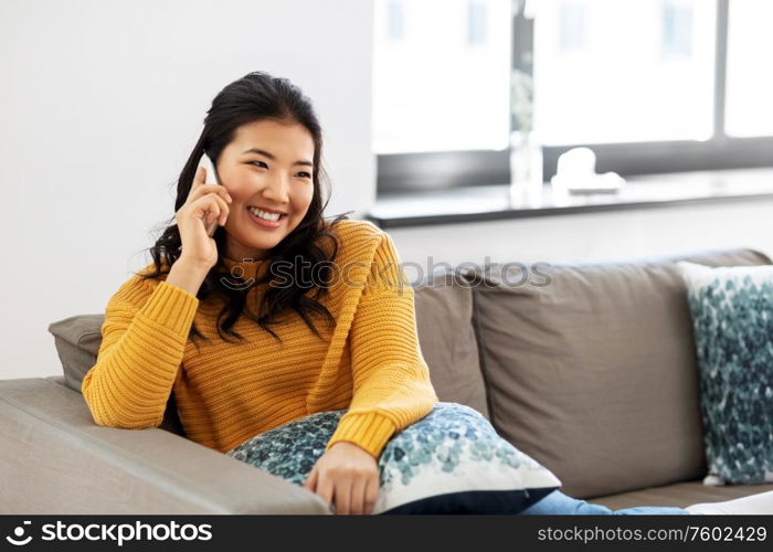 people, technology and communication concept - happy smiling asian young woman in yellow sweater sitting on sofa and calling on smartphone at home. smiling asian woman calling on smartphone at home