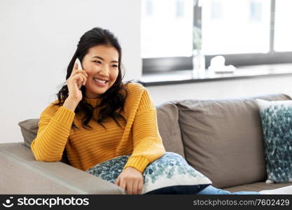 people, technology and communication concept - happy smiling asian young woman in yellow sweater sitting on sofa and calling on smartphone at home. smiling asian woman calling on smartphone at home