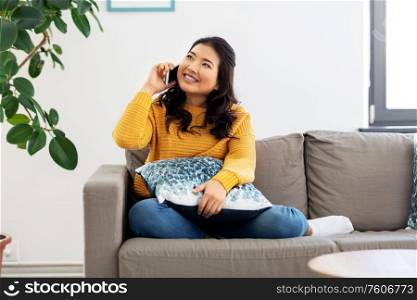 people, technology and communication concept - happy smiling asian young woman in yellow sweater sitting on sofa and calling on smartphone at home. smiling asian woman calling on smartphone at home