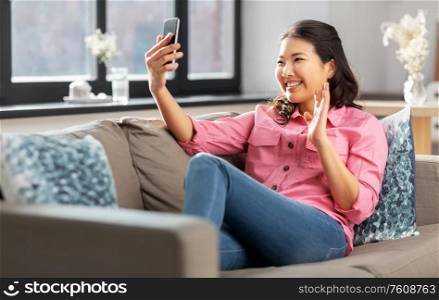 people, technology and communication concept - happy smiling asian young woman in pink shirt sitting on sofa and having video call on smartphone at home. woman with smartphone having video call at home