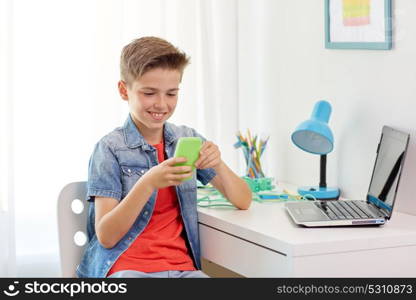 people, technology and communication concept - happy smiling boy with smartphone and laptop computer at home desk. happy boy with smartphone and laptop at home