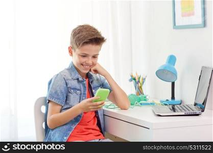 people, technology and communication concept - happy smiling boy with smartphone and laptop computer at home desk. happy boy with smartphone and laptop at home