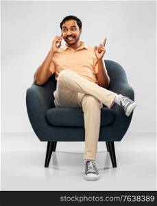 people, technology and communication concept - happy smiling young indian man sitting in chair calling on smartphone over grey background. happy indian man calling on smartphone in chair
