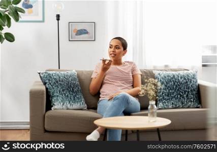 people, technology and communication concept - happy young african american woman using voice command recorder on smartphone at home. woman recording voice with smartphone at home