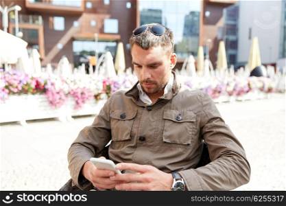 people, technology and communication concept - young man with smartphone texting message at city street cafe terrace