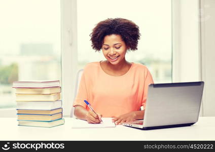 people, technology and education concept - happy african american young woman sitting at table with laptop computer and books at home