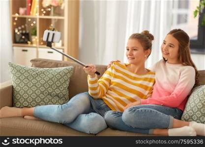 people, technology and friendship concept - happy teenage girls taking picture with smartphone on selfie stick sitting on sofa at home. happy girls taking selfie with smartphone at home
