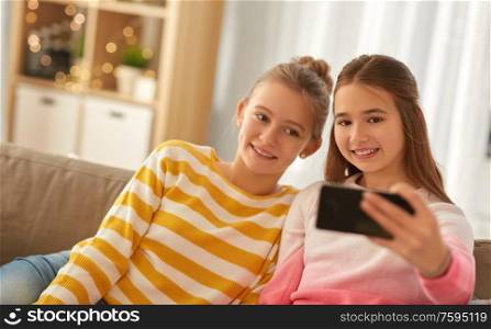 people, technology and friendship concept - happy teenage girls taking selfie with smartphone sitting on sofa at home. happy girls taking selfie with smartphone at home