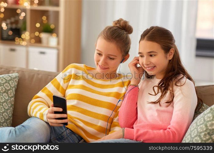 people, technology and friendship concept - happy teenage girls with smartphone and earphones listening to music sitting on sofa at home. happy girls with smartphone and earphones at home