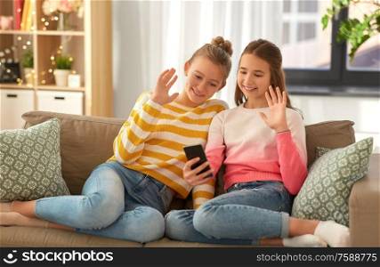 people, technology and friendship concept - happy teenage girls with smartphone sitting on sofa and having video call at home. girls having video call on smartphone at home