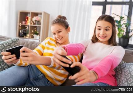 people, technology and friendship concept - happy teenage girls with smartphones sitting on sofa and playing game at home. girls and playing game on smartphones at home