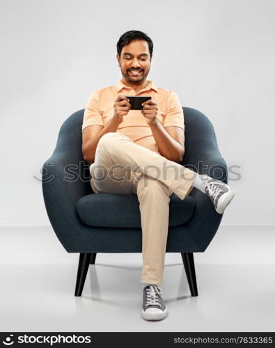 people, technology and furniture concept - happy smiling young indian man playing game on smartphone sitting in chair over grey background. happy young indian man with smartphone in chair