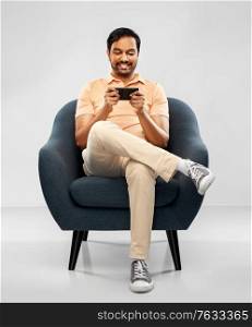 people, technology and furniture concept - happy smiling young indian man playing game on smartphone sitting in chair over grey background. happy young indian man with smartphone in chair