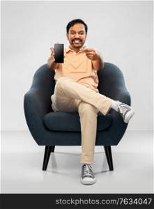 people, technology and furniture concept - happy smiling young indian man showing smartphone sitting in chair over grey background. happy young indian man showing smartphone in chair