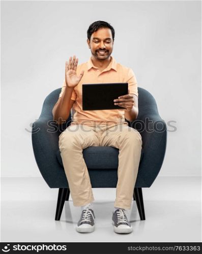 people, technology and furniture concept - happy smiling young indian man with tablet pc computer having video call and waving hand sitting in chair over grey background. indian man with tablet pc having video call