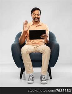 people, technology and furniture concept - happy smiling young indian man with tablet pc computer having video call and waving hand sitting in chair over grey background. indian man with tablet pc having video call
