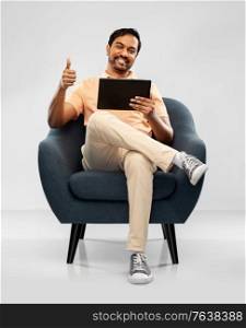 people, technology and furniture concept - happy smiling young indian with tablet pc computer man sitting in chair and showing thumbs up over grey background. happy indian man with tablet pc showing thumbs up