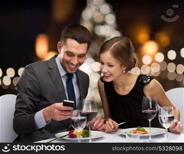 people, technology and holiday concept - smiling couple taking picture of main course with smartphone camera at restaurant over christmas tree background. smiling couple eating main course at restaurant