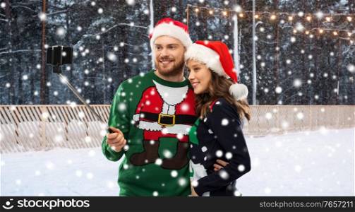 people, technology and holidays concept - happy couple in santa hats and sweaters taking picture with smartphone on selfie stick over skating rink and snow on background. happy couple in christmas sweaters taking selfie