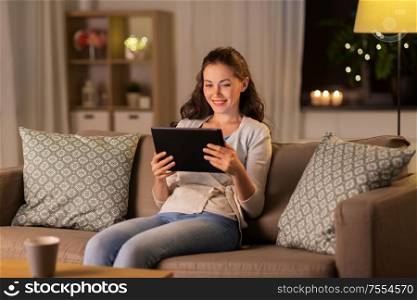 people, technology and internet concept - happy woman sitting on sofa with tablet pc computer at home in evening. happy woman with tablet pc at home in evening