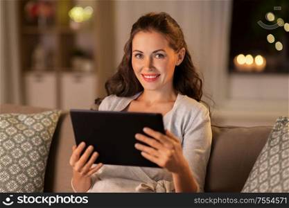 people, technology and internet concept - happy woman sitting on sofa with tablet pc computer at home in evening. happy woman with tablet pc at home in evening