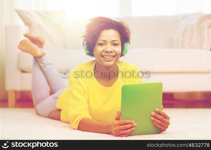 people, technology and leisure concept - happy african american young woman lying on floor with tablet pc computer and headphones listening to music at home
