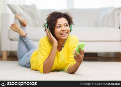 people, technology and leisure concept - happy african american young woman lying on floor with smartphone and headphones listening to music at home