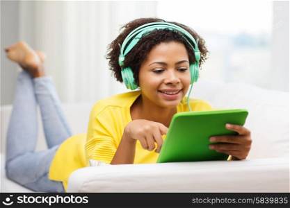 people, technology and leisure concept - happy african american young woman lying on sofa with tablet pc computer and headphones listening to music at home