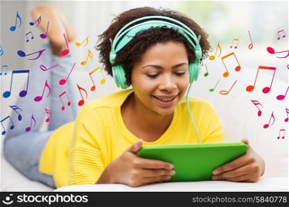 people, technology and leisure concept - happy african american young woman lying on sofa with tablet pc computer and headphones listening to music at home over notes background