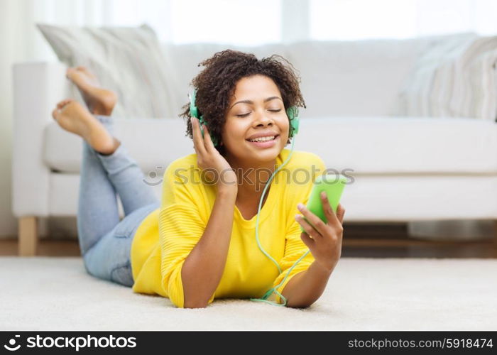 people, technology and leisure concept - happy african american young woman lying on floor with smartphone and headphones listening to music at home