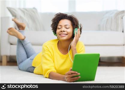 people, technology and leisure concept - happy african american young woman lying on floor with tablet pc computer and headphones listening to music at home