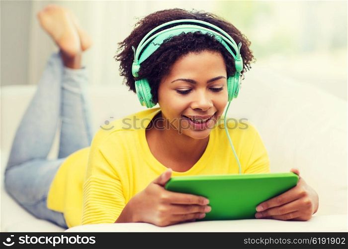 people, technology and leisure concept - happy african american young woman lying on sofa with tablet pc computer and headphones listening to music at home