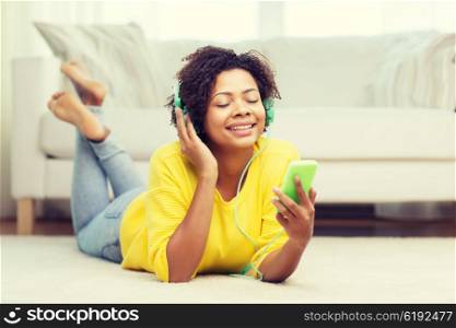 people, technology and leisure concept - happy african american young woman lying on floor with smartphone and headphones listening to music at home
