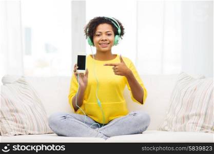 people, technology and leisure concept - happy african american young woman sitting on sofa with smartphone and headphones listening to music at home